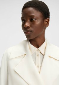 Weißer maßgeschneiderter Blazer mit breiten Revers, getragen über einem hellen Hemd mit Kragen und Knopfdetails. Glattes Material mit strukturierter Passform.