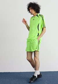 Šviesiai žalia sportinė apranga su juodomis detalėmis; apima trumpas rankoves turintį marškinėlį ir šortus. Modelis dėvi tamsius sportbačius ir baltas kojines.