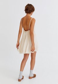 Cremfarbendes ärmelloses Kleid mit niedrigem Rücken, geraffter Taille und fließendem Rock. Verfügt über zarte Spitzenverzierungen und eine sanfte Textur.