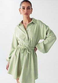 Šviesiai žalia marškinėlių suknelė su apykakle, sagų priekis, elastinė juosmens dalis ir platėjanti sijonas. Ilgos rankovės su surinktomis rankogaliais ir raišteliu.