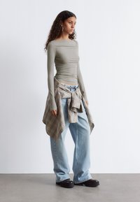 Langärmeliges, olivgrünes Oberteil mit Raffung, kombiniert mit hellblauen, lässigen Jeans und einem karierten Hemd, das um die Taille gebunden ist. Schwarze Plateauschuhe.