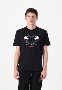 Black short-sleeve t-shirt featuring a graphic design of mirrored dog heads in white, with "HUGO" printed in red below. Smooth cotton material.