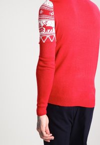 Persona che indossa un maglione rosso con motivi festivi bianchi, tra cui renne sulle maniche, abbinato a pantaloni scuri, in piedi davanti a uno sfondo neutro.