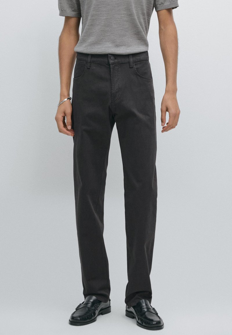 Pantaloni grigi scuri in cotone con design a gamba dritta. Presentano tasche anteriori e chiusura a bottone. Indossati con una camicia grigia e scarpe nere.