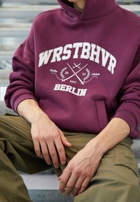 Bordeaux hoodie met witte grafische tekst en details. Voorzien van ribgebreide manchetten en een voorzak. Gecombineerd met olijfgroene broek.