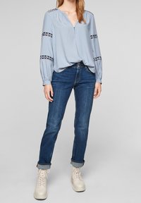 Vaaleansininen pitkähihaisen pusero, jossa on V-pääntie ja nappi, ja mustat pitsikoristeet hihoissa. Yhdistetty tummansinisiin farkkuihin ja beigeihin nilkkureihin.