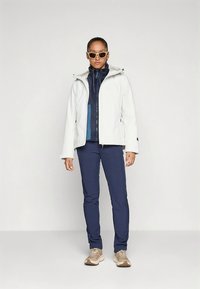 White waterproof jacket with a hood, layered over a navy blue fleece, paired with navy trousers and beige hiking shoes. Clear, minimal design.