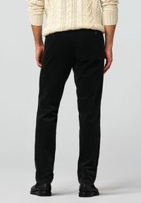 Schwarze Cordhosen mit gerippter Textur, schmaler Passform und Gesäßtaschen. Kombiniert mit einem hellbeigen Strickpullover mit Zopfmuster und schwarzen Schuhen.