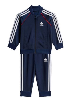 Marineblå treningsdrakt laget av glatt stoff, med hvite tre-stripe detaljer, full glidelåsjakke og elastiske mansjetter og midjebånd. Adidas-logo på forsiden.