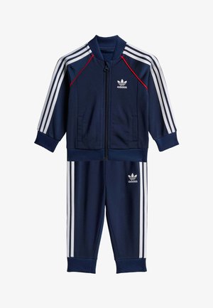fato de treino azul marinho feito de tecido suave, apresentando detalhes de riscas brancas, casaco com fecho éclair completo e punhos e cintura elásticos. Logotipo da Adidas na parte da frente.