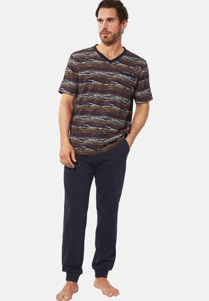 Mann trägt ein kurzärmliges, mehrfarbig gestreiftes V-Ausschnitt-Shirt und marineblaue Jogginghosen, steht barfuß mit einer Hand in der Tasche.