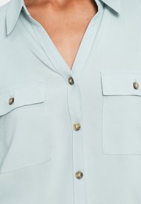 Jasnoniebieska koszula z kołnierzykiem, z dwoma kieszeniami na piersi i okrągłymi, kontrastującymi guzikami; gładka tekstura tkaniny.