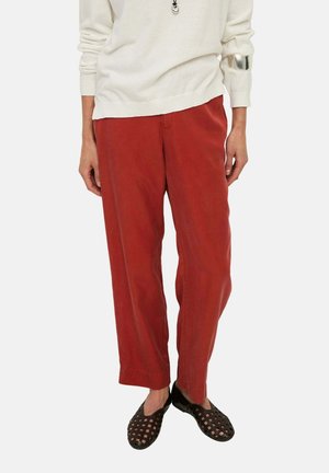 Persona con pantalones rojos holgados, camiseta blanca de manga larga, pulsera plateada en el brazo izquierdo y zapatos negros tejidos sin cordones.