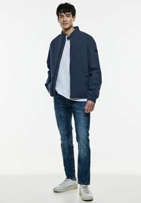 Chaqueta bomber navy, camiseta blanca, jeans azules y zapatillas blancas. La chaqueta cuenta con un collarín tipo stand y cierre de cremallera. Diseño simple.