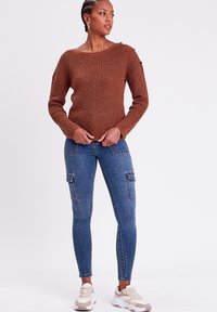 Pull en tricot marron avec un motif texturé, décolleté large et accents en boutons. Associé à un jean cargo bleu ajusté et des baskets claires.
