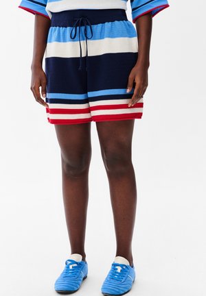 Nogi w granatowych, niebieskich, białych i czerwonych pasiastych sweterkowych szortach, w połączeniu z pasującymi niebieskimi trampkami i białymi skarpetkami na białym tle.