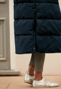 Manteau long matelassé bleu marine avec boutons pression, associé à des leggings texturés olive et des chaussures de sport argentées, présentant des détails contrastants.