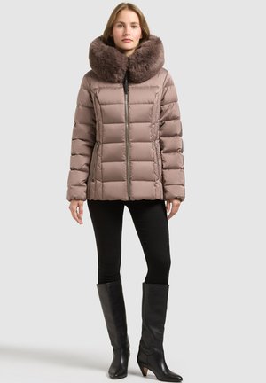Femme portant une doudoune taupe avec un large col en fourrure, des leggings noirs et des bottes noires à talons jusqu'aux genoux, debout devant un fond uni.