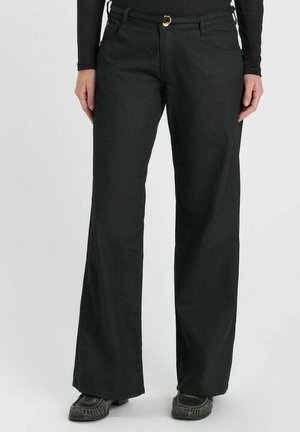 Femme portant un pantalon noir à jambes larges avec une fine ceinture et des chaussures noires, debout devant un fond clair uni.