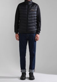 Zwarte puffervest met een rits aan de voorkant en hoge kraag, gedragen over een zwart langemouwenshirt. Gepaard met blauwe jeans en zwarte schoenen.