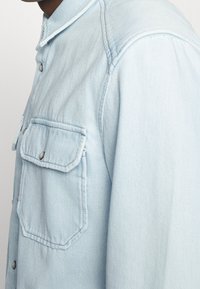 Camicia di jeans azzurro chiaro con colletto strutturato, bottoni a pressione e due tasche frontali. Il tessuto ha un aspetto morbido e strutturato.