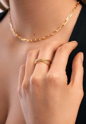 Goldring mit verflochtenen Bändern und geflochtenem Design, getragen an einer Hand mit glatter Hauttextur. Sichtbare Goldkette.
