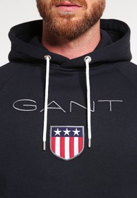 Marineblauer Kapuzenpullover mit weißen Kordeln, verziert mit dem "GANT"-Logo, das über einem Schild mit roten und weißen Streifen und Sternen aufgestickt ist.