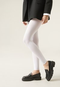 Hvide leggings med en glat tekstur, parret med sorte platform loafers og en sort blazer, vises mod en lys baggrund.