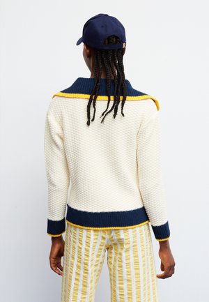 Persona con largas trenzas que lleva un jersey blanco texturizado con ribetes azul marino y amarillo, pantalones a rayas amarillas y blancas y una gorra azul marino, de espaldas.