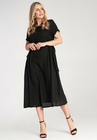 Czarna, zwiewna midi sukienka z krótkimi rękawami, z kieszeniami na sznurek po bokach i lekką fakturą. Noszona z czarnymi sandałami na obcasie.