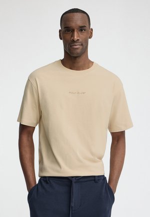 Man staat met handen in de zakken, draagt een beige korte mouwen shirt met de tekst "POLO CLUB" en donkerblauwe broek op een effen achtergrond.