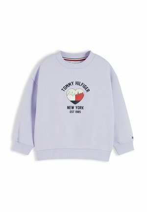 Jong meisje met lang donker haar, gekleed in een lichtpaarse Tommy Hilfiger trui en een blauwe spijkerbroek, staand voor een effen witte achtergrond.