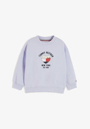Jeune fille aux longs cheveux foncés portant un sweat-shirt Tommy Hilfiger violet clair et un jean bleu, debout devant un fond blanc uni.