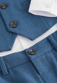 Tissu de costume texturé bleu avec pantalon assorti et col de chemise blanc. Boutons noirs avec détail de marque "Baker".