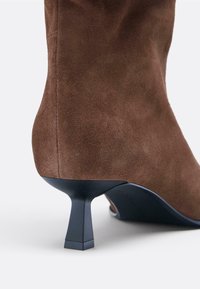 Bottine en daim marron avec un talon bleu effilé, présentant une texture lisse et un design élégant, mettant en valeur des détails de coutures subtiles.