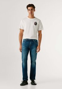 Joven de pie con jeans azules oscuros, camiseta blanca con un pequeño rosa y logo de texto, y zapatos negros con cordones, contra un fondo liso.