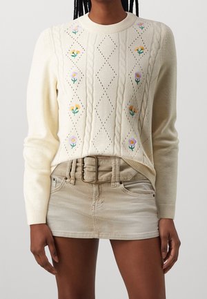 Pull en maille crème avec broderie florale, associé à une mini-jupe beige en velours côtelé et une large boucle de ceinture.