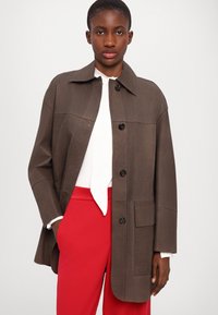 Jachetă oversized de culoare maro, cu guler și nasturi pe partea din față, și buzunare mari. Pur și simplu, peste un top alb și pantaloni roșii. Textură fină.