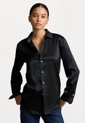 Camicia nera di satin con bottoni, maniche lunghe, colletto pieghettato e dettagli in bottone bianchi, abbinata a jeans blu scuro. Texture liscia.