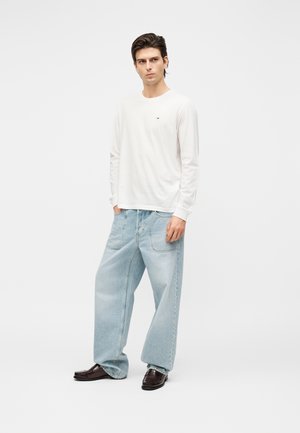 Νέος άνδρας με σκούρα μαλλιά, φοράει λευκό πουκάμισο με μακριά μανίκια, ανοιχτό μπλε τζιν παντελόνι με φαρδιά μπατζάκια και σκουριά καφέ παπούτσια, στέκεται με τα χέρια στις τσέπες.