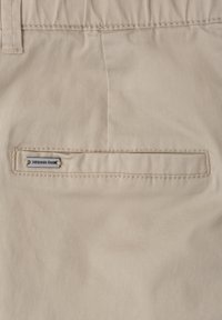Poche arrière d’un pantalon beige avec fente horizontale et petite étiquette en métal portant la mention « Street One » sur la bordure cousue.