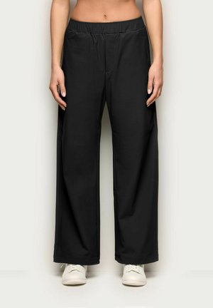 Person trägt schwarze Hosen mit weitem Bein und elastischem Bund sowie weiße Sneakers, steht vor einem neutralen Hintergrund.