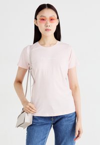 T-shirt rosa claro de manga curta feita de um tecido suave, com um logo discreto em branco. Estilizado com jeans azuis e uma pequena bolsa cinza.
