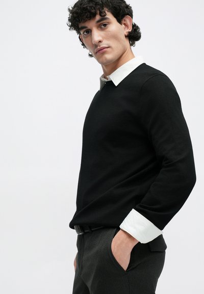 Pull noir avec col blanc, manches longues et coupe décontractée. Pantalons gris avec tissu texturé. Le modèle est posé avec les mains dans les poches.