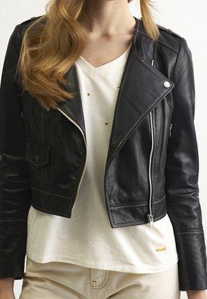 Zwarte cropped leren jas met zilveren ritsen, gedragen over een wit V-hals shirt met gouden studs en beige broek.