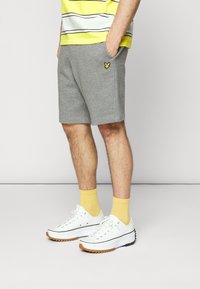 Grå bomullsshorts med sidelommer og en liten gul logo. Kombinert med hvite joggesko med svart og gummisåle og gule sokker.