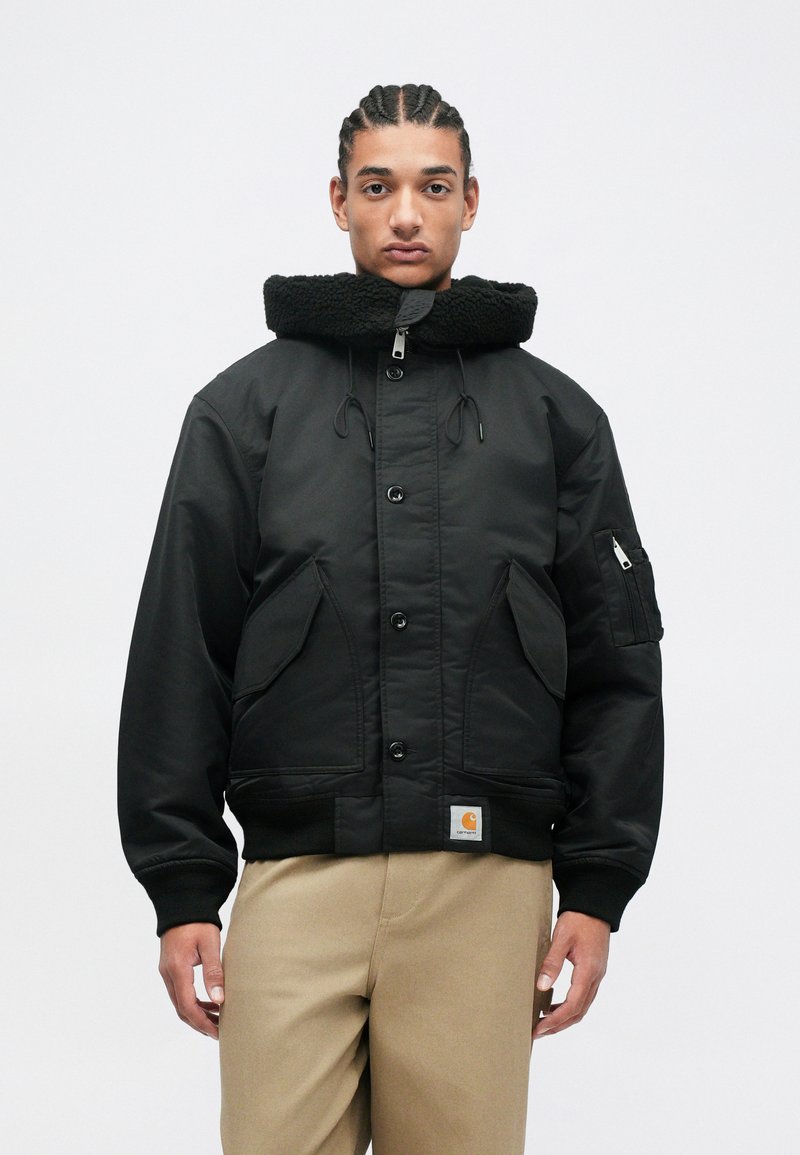 Veste bomber isolante noire avec un col haut en fourrure, de grandes poches avant, et des poignets et un ourlet côtelés. Présente un patch logo.