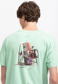 T-shirt en coton vert menthe avec un imprimé graphique coloré au dos d'un ancien appareil photo entouré de planches en bois.