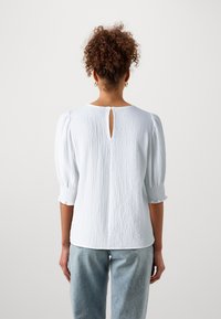 Camicetta bianca con texture e maniche a sbuffo, caratterizzata da un'apertura a chiave sul retro e polsini elasticizzati. Abbinata a jeans in denim chiaro.