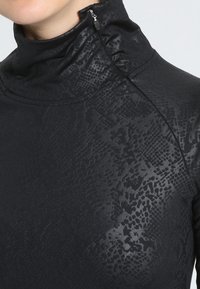 Acercamiento de un cuello de tortuga negro con un sutil patrón de piel de serpiente y un pequeño detalle de cremallera cerca del cuello.